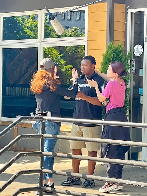 “Too Southern"?! Julia Fowler Would Like a Word - 6 Three people are filming a scene outside a building; one, possibly Julia Fowler of Too Southern, holds up her hand, another faces her, and a boom microphone is visible above them.