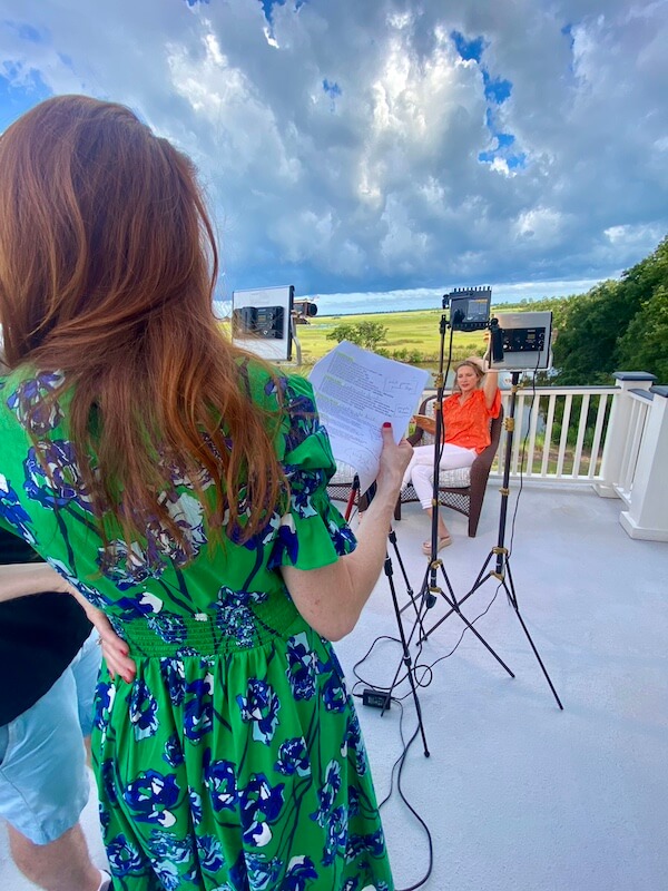 “Too Southern"?! Julia Fowler Would Like a Word - 5 Julia Fowler, creator of Too Southern, holds papers in a green dress while another woman sits before cameras and lights on an outdoor terrace with a scenic landscape in the background.