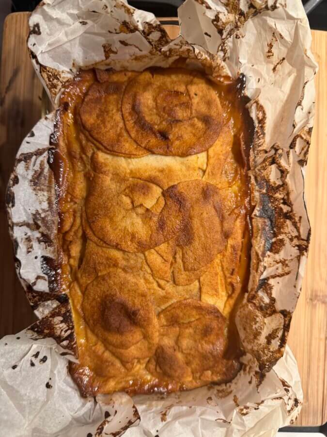 RECIPE: Invisible French Apple Cake - 4 A baked dish lined with parchment paper, featuring browned, layered slices on top—reminiscent of an Invisible French Apple Cake—rests on a wooden surface, tempting with its rustic charm.
