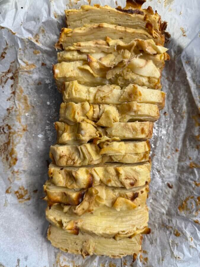 RECIPE: Invisible French Apple Cake - 5 A sliced Apple Ribbon Loaf rests on crumpled parchment paper, showing visible layers of thinly cut apples and a golden-brown top.