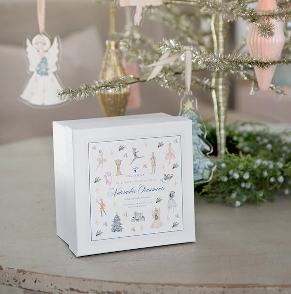 14 Southern Ornament & Stocking Shops We Love - 9 A white box labeled "Nutcracker Ornaments" sits on a table near a decorated Christmas tree adorned with angel, pastel, and southern ornaments.