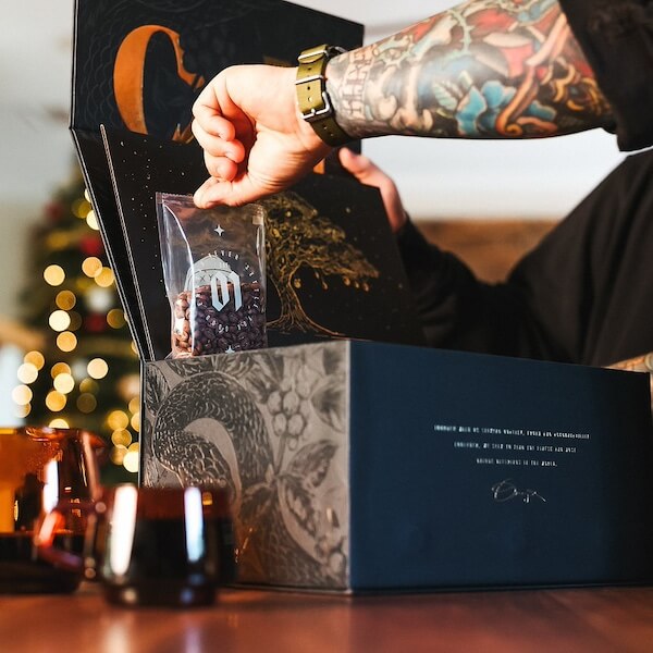 The BEST Advent Calendars for 2025 - 11 A tattooed person opens a decorative box, reminiscent of advent calendars, and holds a small bag of coffee beans, with candles and a blurred Christmas tree in the background.