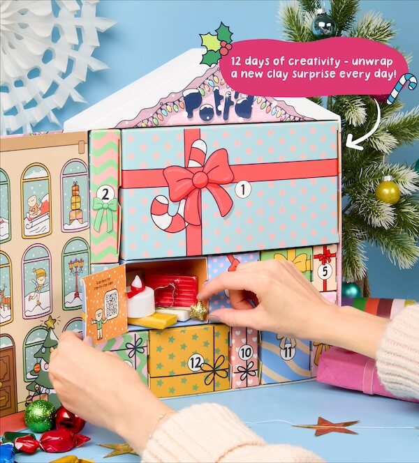 The BEST Advent Calendars for 2025 - 24 A person opens a compartment in a festive advent calendar with a holiday theme, revealing small clay crafting items inside. A sign reads, "12 days of creativity – unwrap a new clay surprise each day with our unique advent calendars!"