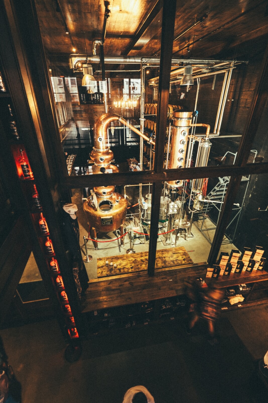Discovering the heart of Nashville, this distillery interior showcases copper stills and pipes behind glass walls, with bottles of local favorites displayed along the side and warm wooden floors throughout the space.