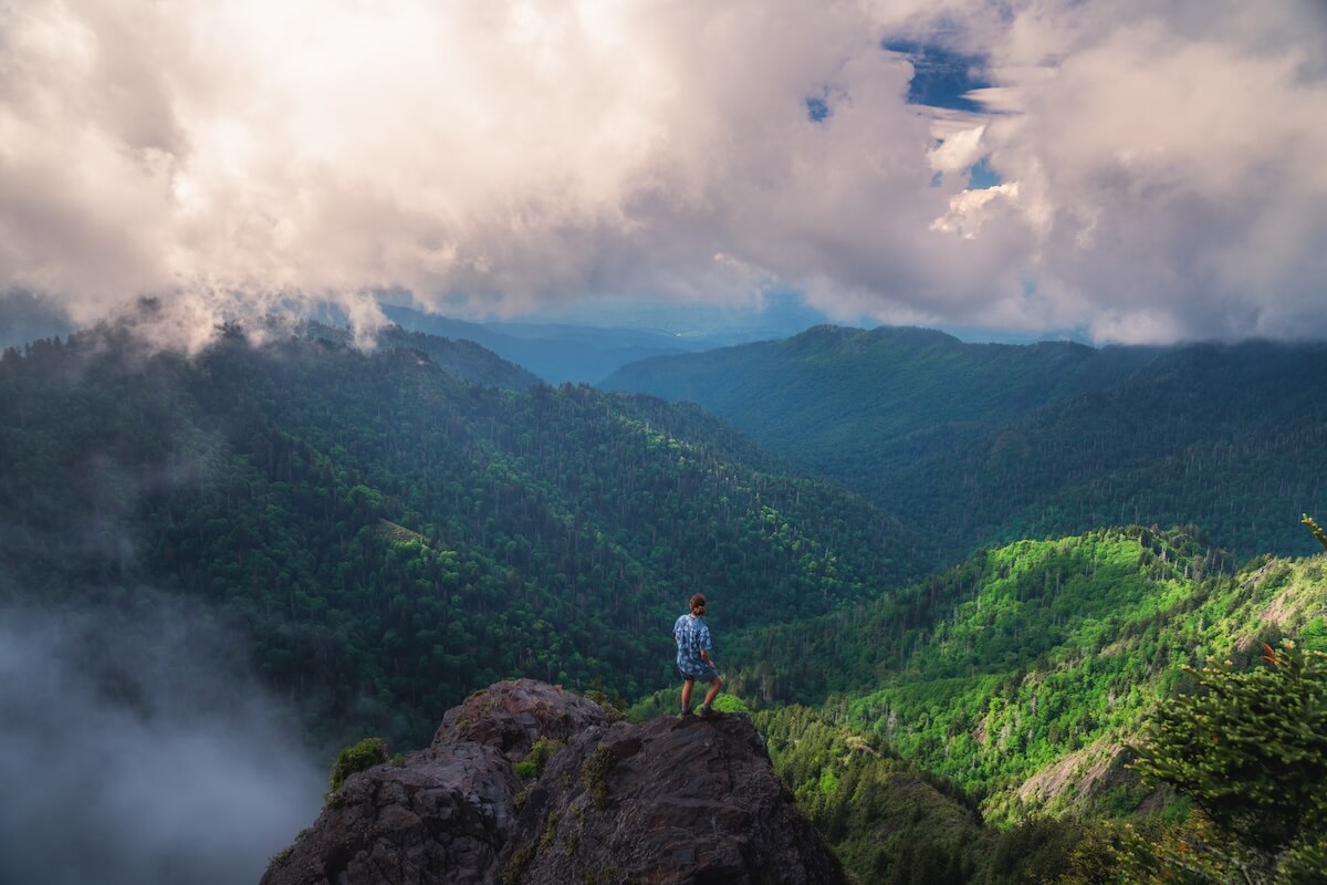 A person stands on a rocky outcrop overlooking a lush, green mountainous landscape—perfect for those seeking Tennessee outdoor escapes, with clouds and sunlight illuminating the adventure that awaits.