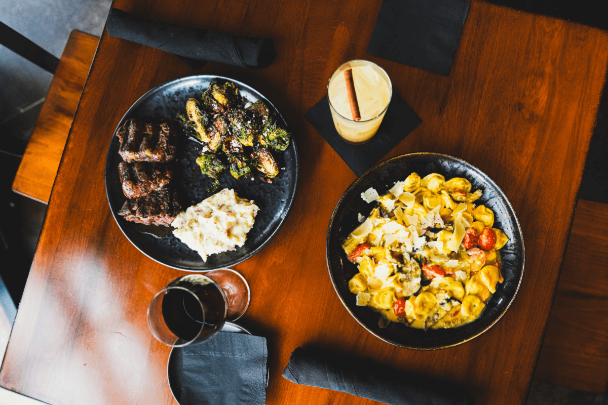 5 Beloved Tennessee Destinations for Adventure Lovers - 10 Two plates of food on a wooden table—grilled meat with Brussels sprouts and potato salad, plus pasta and veggies—set the scene for memorable Tennessee adventures, complemented by glasses of wine and an iced drink.