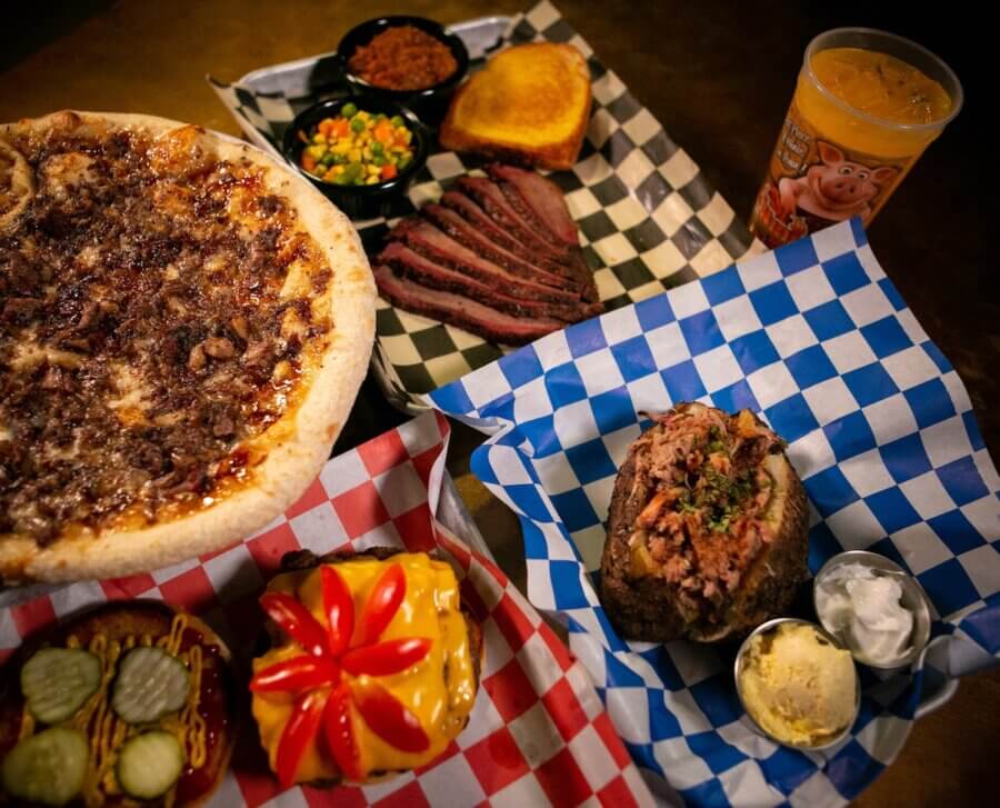 Culinary Road Trip: 4 Tennessee Towns for Food Lovers - 12 A table with Tennessee food favorites: barbecue pizza, smoked brisket slices with sides, a cheeseburger stacked with pickles and tomato, a baked potato loaded with toppings, and a cup of juice.