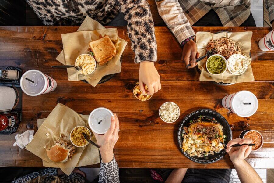 Culinary Road Trip: 4 Tennessee Towns for Food Lovers - 13 Four people sit around a wooden table enjoying barbecue-style Tennessee food with sides, drinks, and sandwiches, viewed from above.