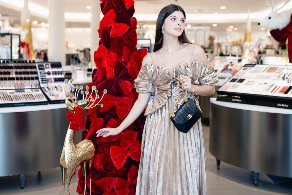 A woman in a metallic off-shoulder dress stands by a gold reindeer statue and red floral decorations in a cosmetics store, showcasing chic holiday looks and holding a black handbag.