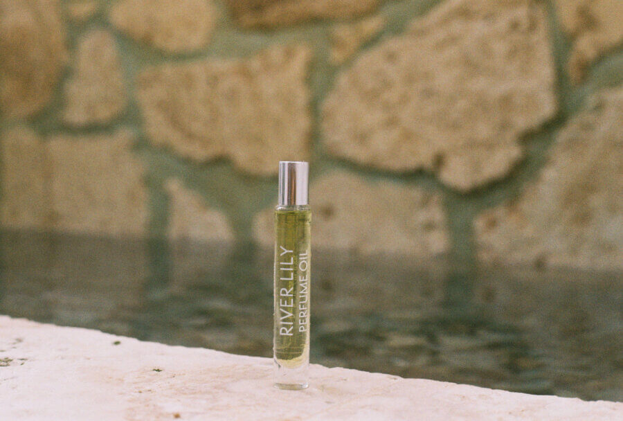 50 Under $50: Your BHAM Holiday Gift Guide - 2 A glass bottle labeled "River Lily Dreaming Oil" stands upright on a stone surface, with a blurry stone wall and water in the background—a serene choice for holiday gifts or those seeking unique gifts under $50.