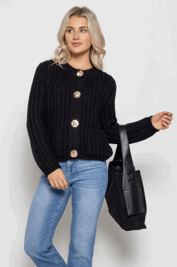 50 Under $50: Your BHAM Holiday Gift Guide - 13 A woman wearing a black knit cardigan with large gold buttons, blue jeans, and holding a black tote bag stands against a plain background.