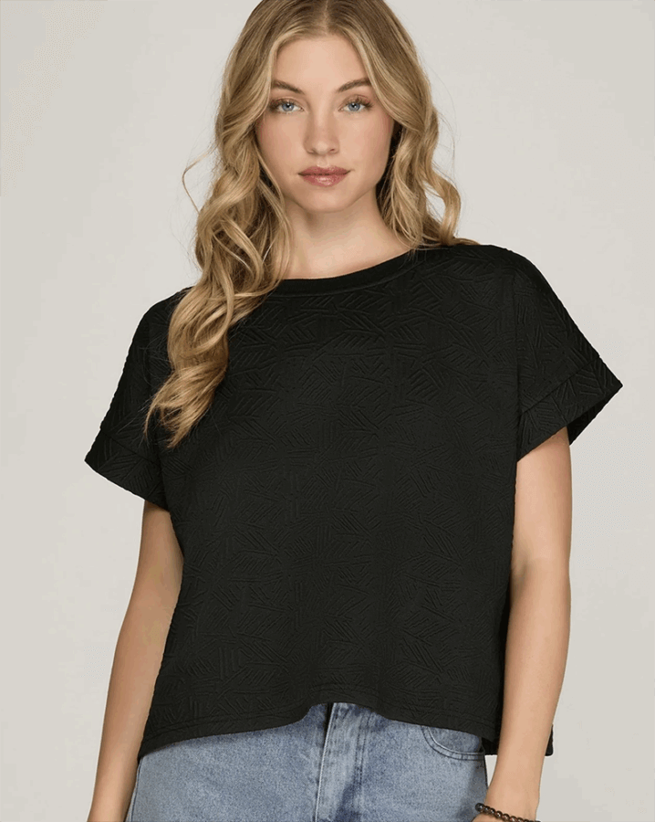 50 Under $50: Your BHAM Holiday Gift Guide - 18 A woman with long wavy blonde hair wearing a loose black short-sleeve textured top and light blue jeans stands against a plain light background, inspiring effortless style for your 2025 Holiday Gift Guide.