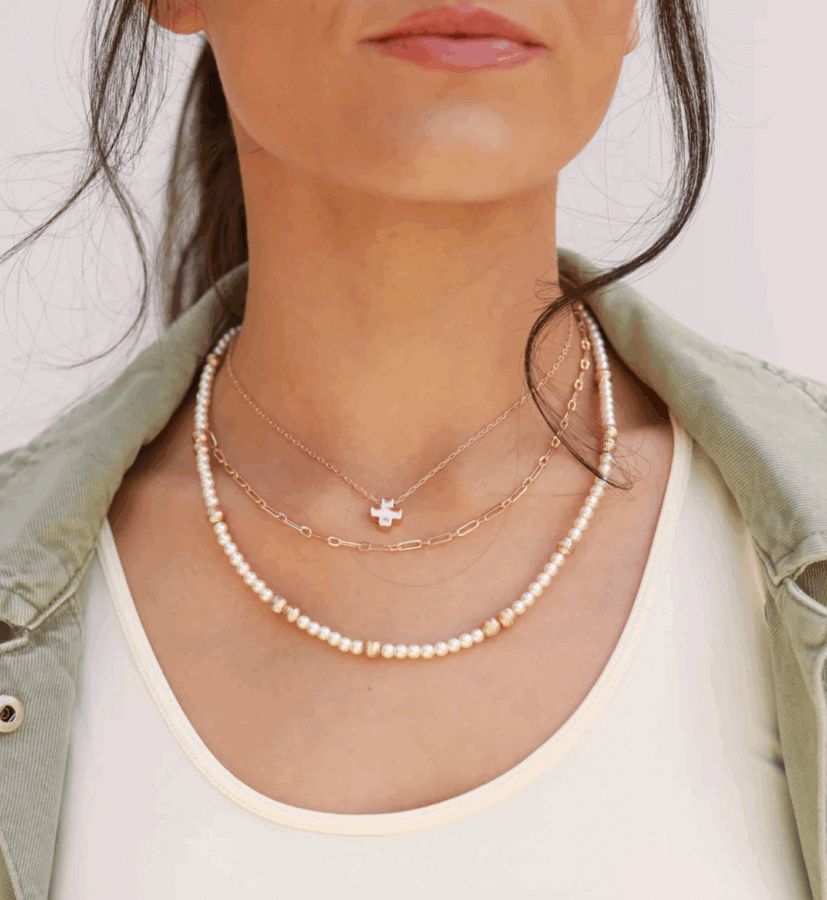 50 Under $50: Your BHAM Holiday Gift Guide - 20 A woman wearing three layered necklaces—one with a cross pendant, one with small chain links, and one with pearls—over a white top and green jacket. Stylish accessories perfect for holiday gifts or finds from the Birmingham Holiday Gift Guide.