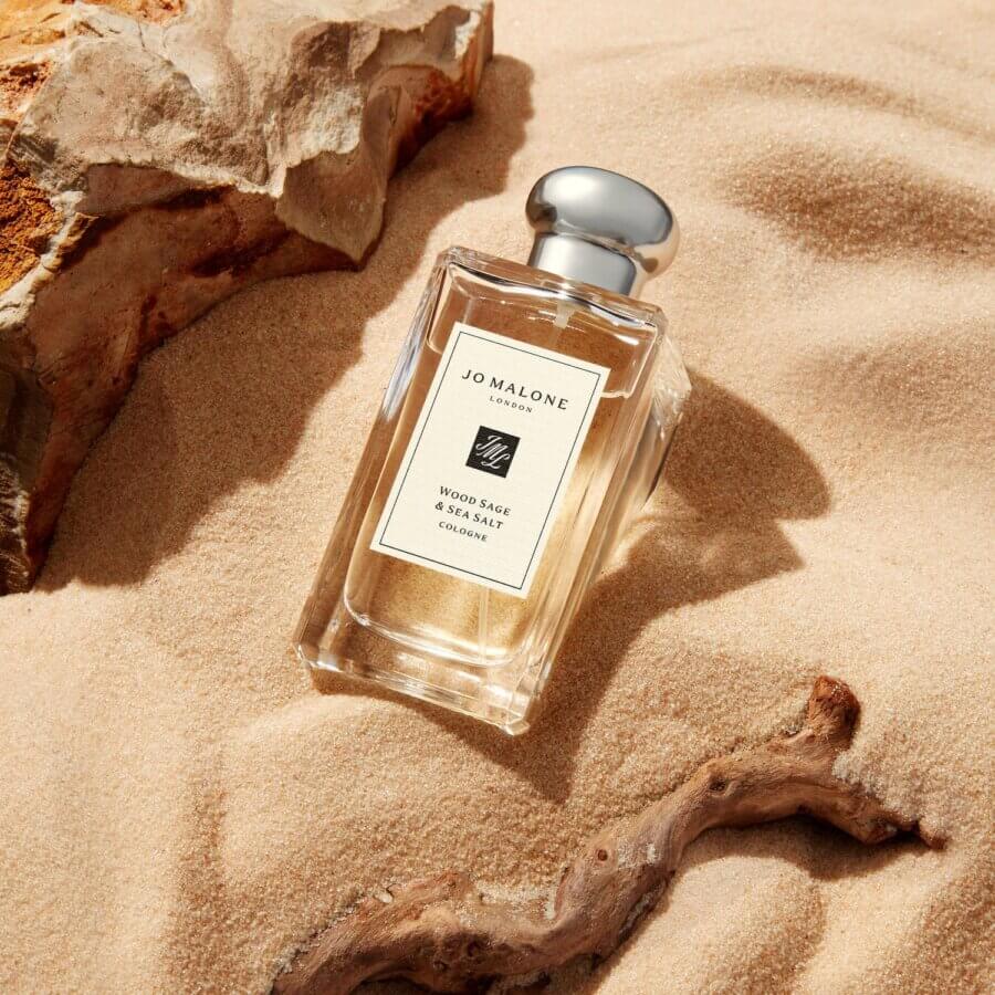 19 Luxurious Birmingham FINDS for the Holidays - 10 A bottle of Jo Malone Wood Sage & Sea Salt cologne rests on sand beside a rock and a piece of driftwood.