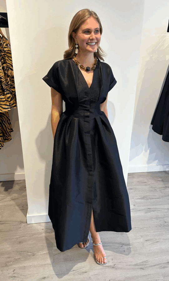 19 Luxurious Birmingham FINDS for the Holidays - 7 A woman stands indoors wearing a black, short-sleeved, ankle-length dress with a flared skirt. She is barefoot and smiling, with her hands in the dress pockets.