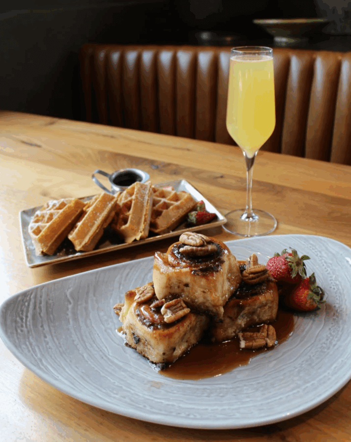 A plate of cinnamon rolls topped with pecans and syrup, fresh strawberries, a side of waffles with syrup, and a glass of orange mimosa on a wooden table—perfect for New Year's Day brunch at your favorite Birmingham restaurants.