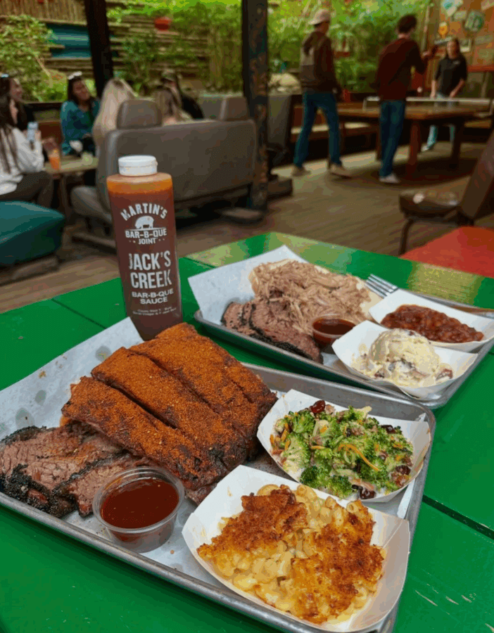 Plates of barbecue with ribs, brisket, pulled pork, sides, and a barbecue sauce bottle on a green table at one of Birmingham Restaurants; people sitting and standing in the background on New Year's Day.