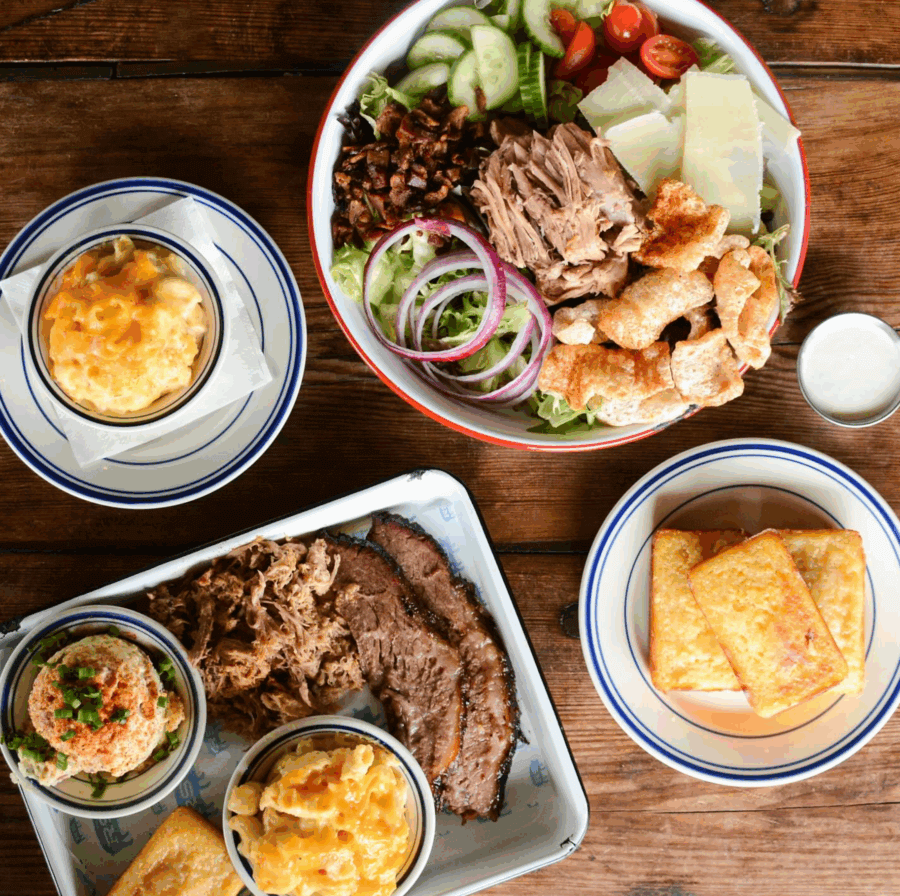 Assorted Southern-style dishes on a wooden table, including a salad with pork, macaroni and cheese, cornbread, brisket, pulled pork, hummus, and a side of dressing—perfect for celebrating New Year's Day at top Birmingham restaurants.