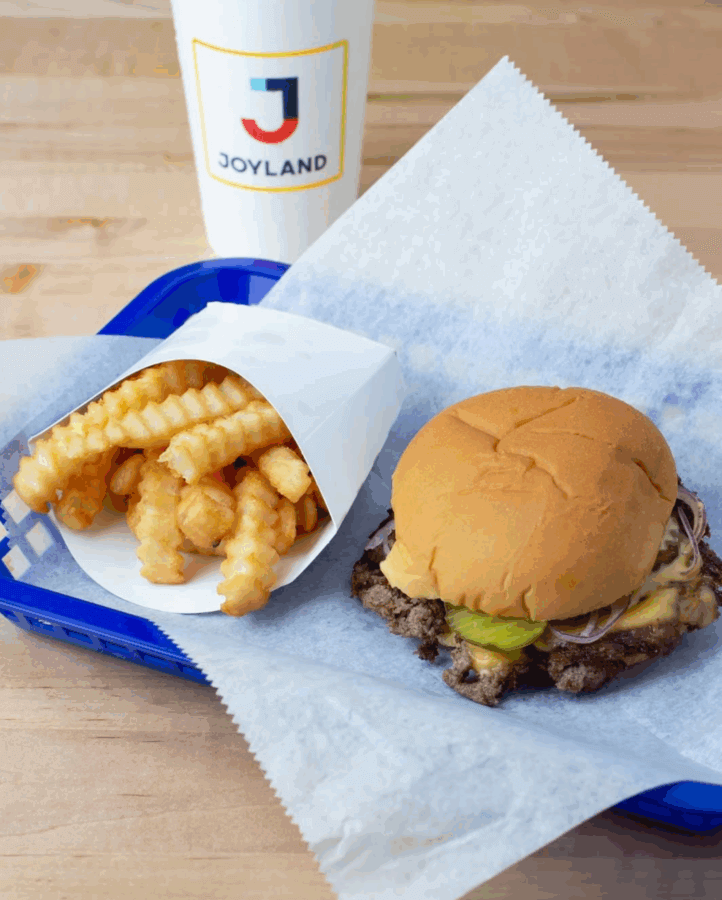 A cheeseburger with pickles and onions, crinkle-cut fries, and a drink in a Joyland cup on a blue tray with parchment paper—perfect for enjoying at open Birmingham restaurants this New Year's Day.