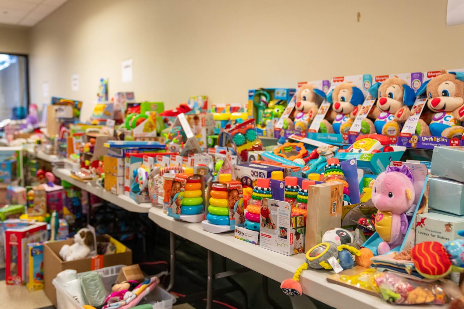 Her Toy Drives Bring Joy & Relief to BHAM Families - 7 Long tables filled with colorful children's toys—including boxed plush animals, stacking rings, and activity sets—are arranged indoors against a beige wall at the Birmingham FACES Circle of Love toy drive organized by Doris Phillips.