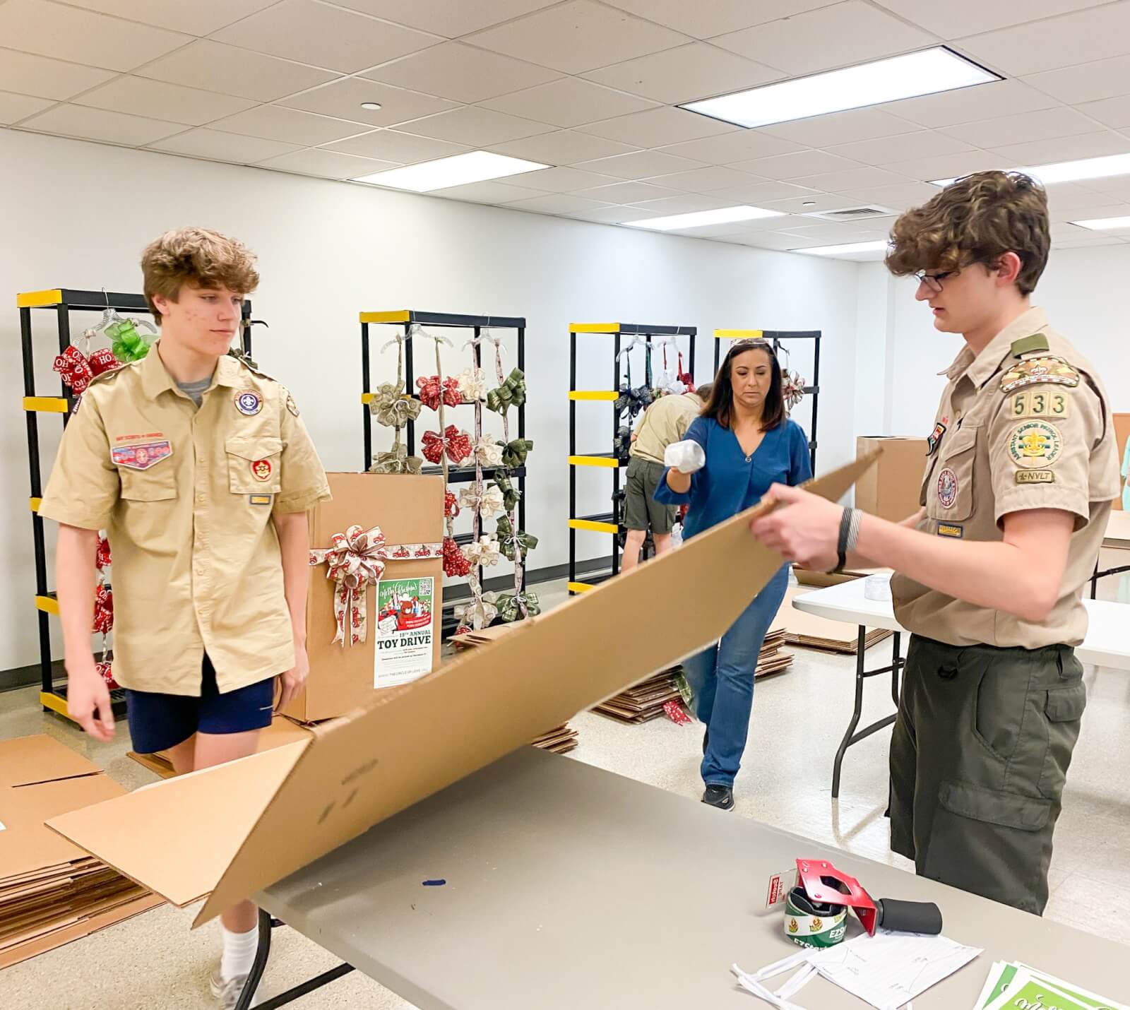 Her Toy Drives Bring Joy & Relief to BHAM Families - 8 Two Boy Scouts assemble cardboard boxes at a table while a woman stands nearby in a room with metal shelves holding wreaths and ribbons for the Circle of Love project.