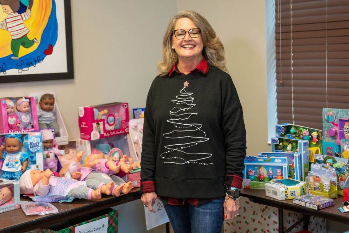 A woman wearing glasses and a black holiday sweater stands in front of tables filled with boxed dolls and children’s toys, supporting Birmingham Families during local Holiday Toy Drives.