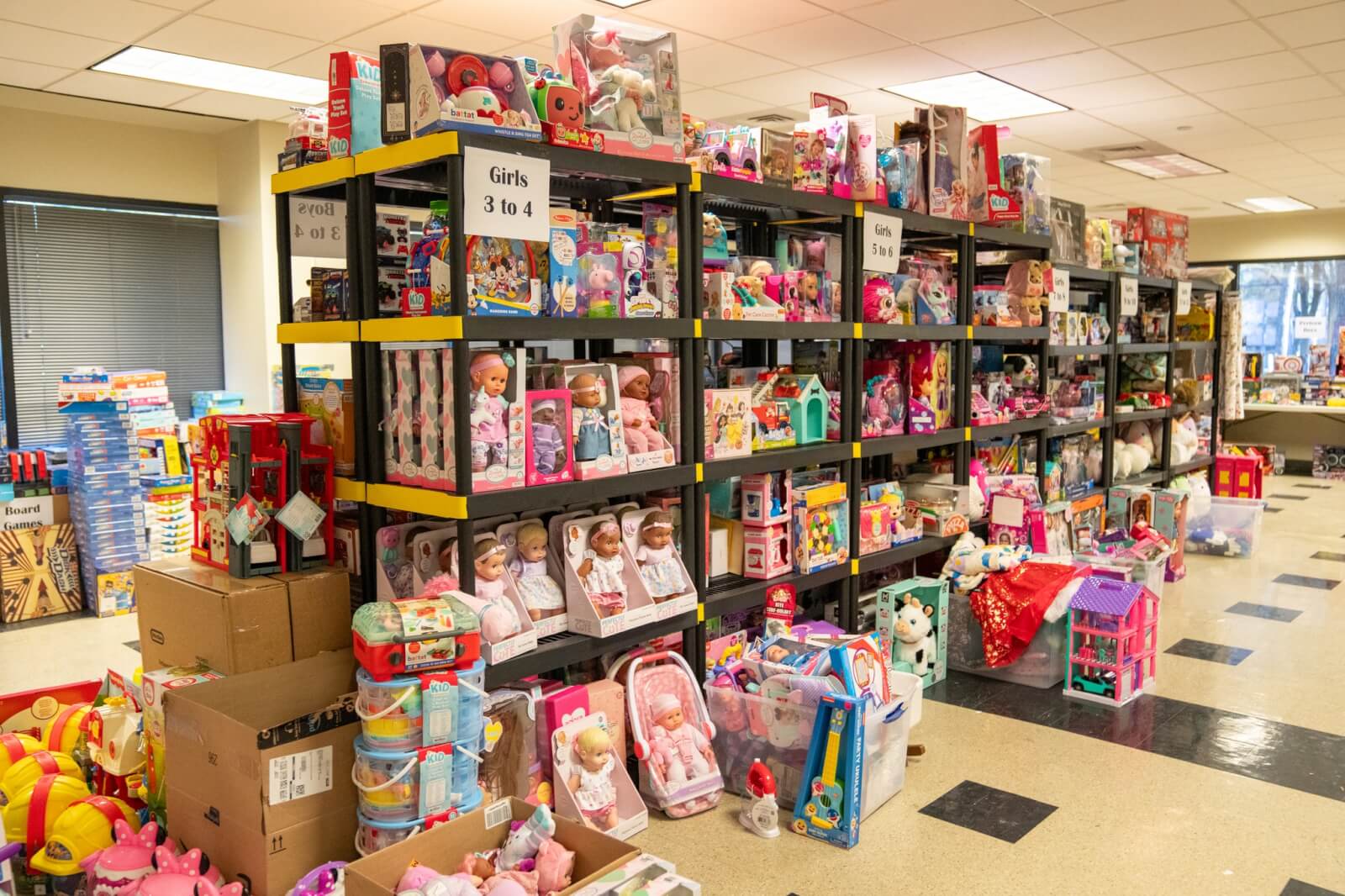 Her Toy Drives Bring Joy & Relief to BHAM Families - 3 Shelves and tables are filled with a variety of boxed toys, dolls, and games, with signs indicating sections for girls ages 3 to 4.