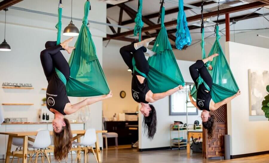 Three women practice aerial yoga, hanging upside down in green hammocks, inside a modern Birmingham studio with exposed ceilings and wooden floors—perfect for unique Birmingham experiences or creative gift ideas.
