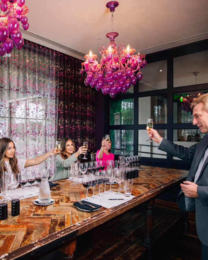 A group of people seated at a wooden table raise glasses in a toast under purple chandeliers, celebrating Birmingham experiences in a modern dining room.