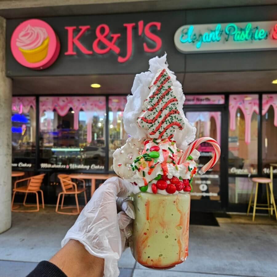 Meet Kristal Bryant of K&J’s Elegant Pastries & Creamery - 5 A gloved hand holds a holiday-themed milkshake topped with whipped cream, a candy cane, berries, and a decorated cookie, in front of K&J's bakery storefront.