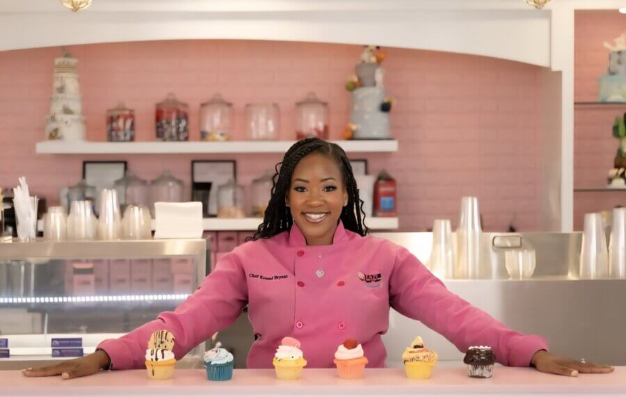 Meet Kristal Bryant of K&J’s Elegant Pastries & Creamery - 2 Kristal Bryant in a pink chef coat stands behind a counter displaying an assortment of colorful cupcakes in a bakery with pink decor at K&J's Elegant Pastries and Creamery.