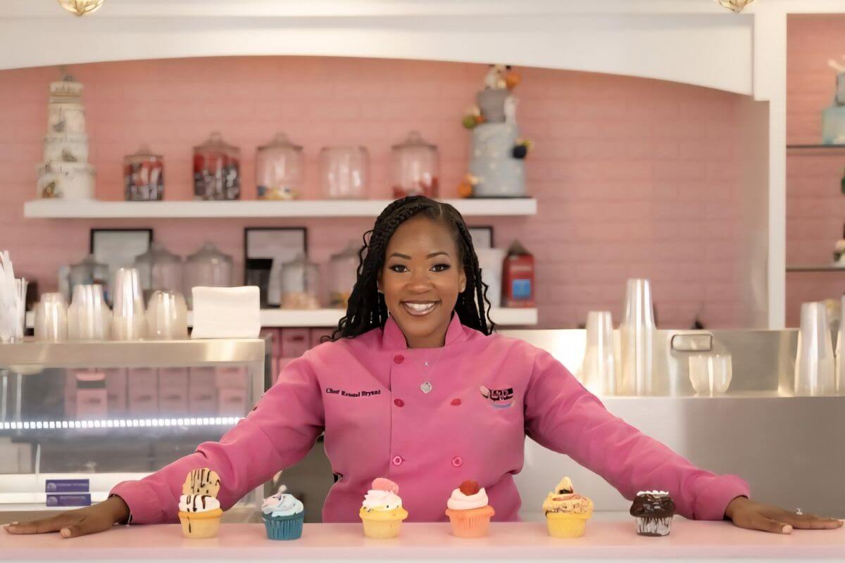 Designer Chef Kristal Bryant stands in a pink coat behind a counter with five elegantly decorated cupcakes at K&J's Elegant Pastries and Creamery.