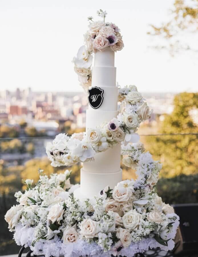 Meet Kristal Bryant of K&J’s Elegant Pastries & Creamery - 6 Tall white wedding cake with five tiers, decorated with cascading white and blush flowers, set outdoors with the Birmingham cityscape in the background.