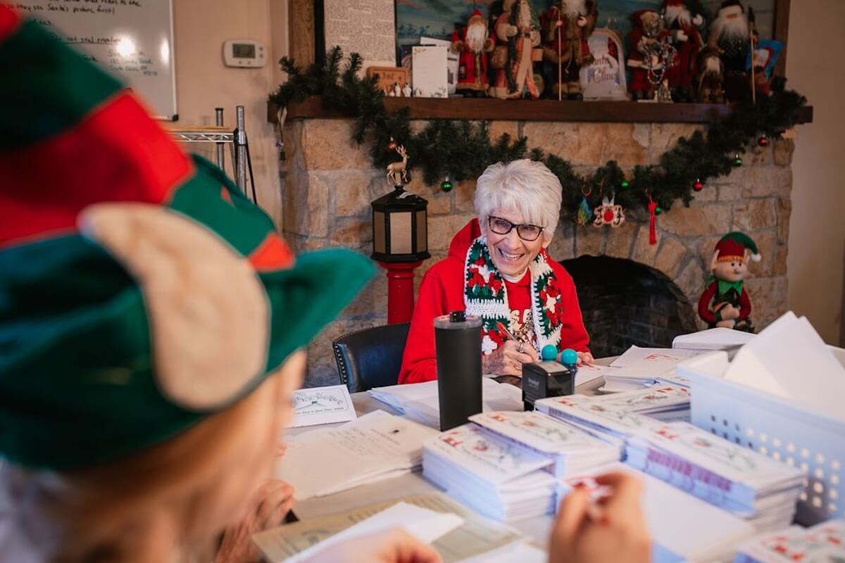 An elderly woman wearing glasses and festive attire sits at a table with mail and papers, smiling, with holiday decorations and a fireplace in the background—just like Pat Koch, Chief Elf of Santa Claus, Indiana.