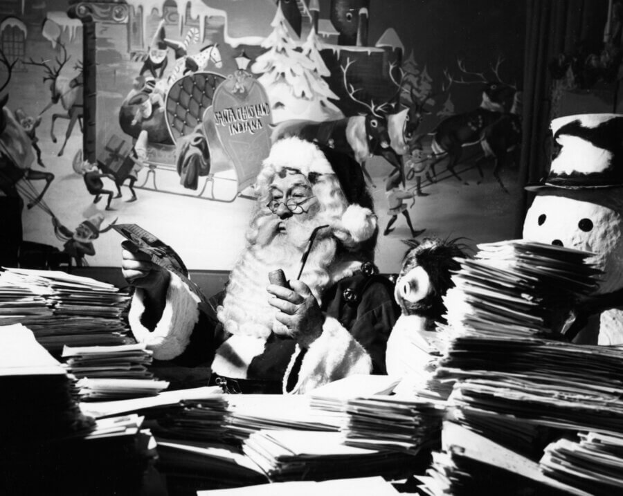 Meet the 94-Year-Old 'Chief Elf' Keeping the Magic of Christmas Alive - 3 Jim Yellig sits behind stacks of letters, holding some in hand, with a snowman figure beside him and a festive mural in the background.