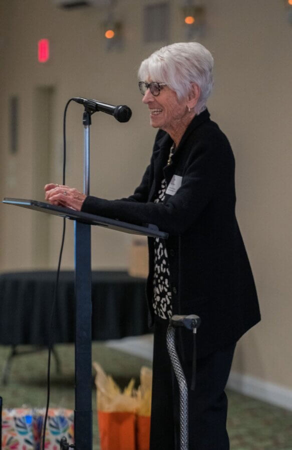 Meet the 94-Year-Old 'Chief Elf' Keeping the Magic of Christmas Alive - 4 Pat Koch, an older woman with short white hair, stands at a podium speaking into a microphone. She wears glasses, a dark jacket, and has a cane beside her. Gift bags are visible in the background.