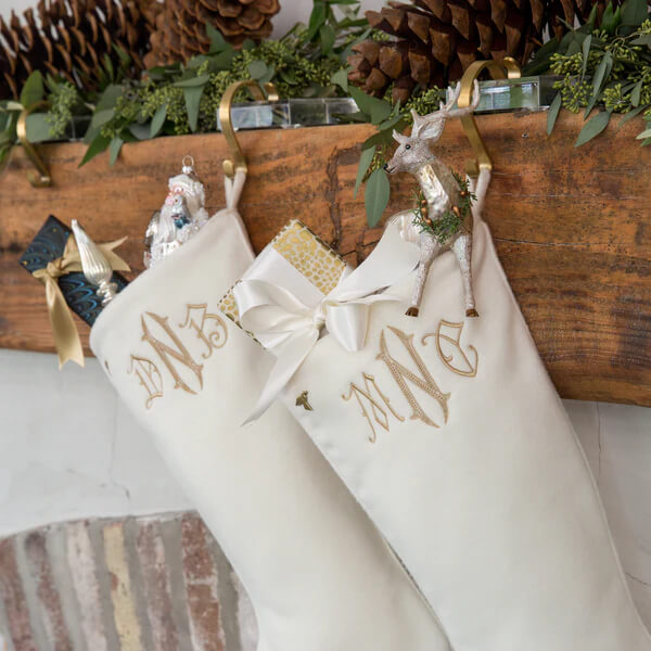 14 Southern Ornament & Stocking Shops We Love - 10 Two white Christmas stockings with gold monograms hang from a mantle adorned with pinecones, greenery, and southern ornaments; one stocking holds a gift and a deer figurine.