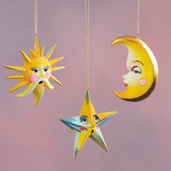 14 Southern Ornament & Stocking Shops We Love - 4 Three southern ornamentsβa sun, star, and crescent moonβeach with painted human-like faces, hang gracefully against a soft pink background.