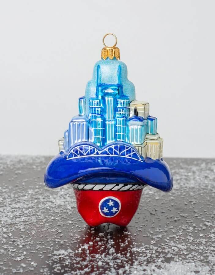 23 Luxurious Nashville FINDS for Holiday 2025 - 15 A luxe Christmas ornament featuring a stylized blue Nashville skyline atop a wide-brimmed hat with a red base displaying the Tennessee state flag emblem, set on a surface with artificial snow—perfect for your holiday décor.