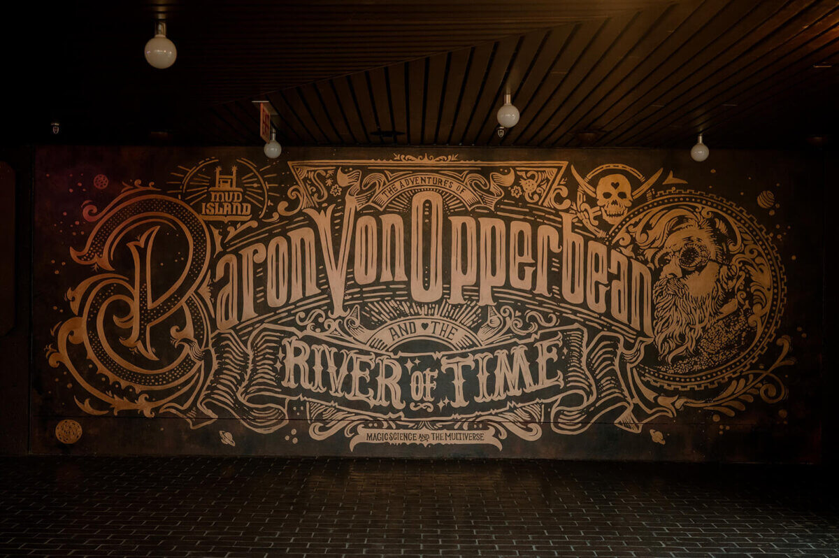 A mural for all ages featuring ornate text that reads "Baron Von Opperbean and the River of Time," with decorative designs and an illustrated bearded figure on the right, capturing a sense of timeless adventure.