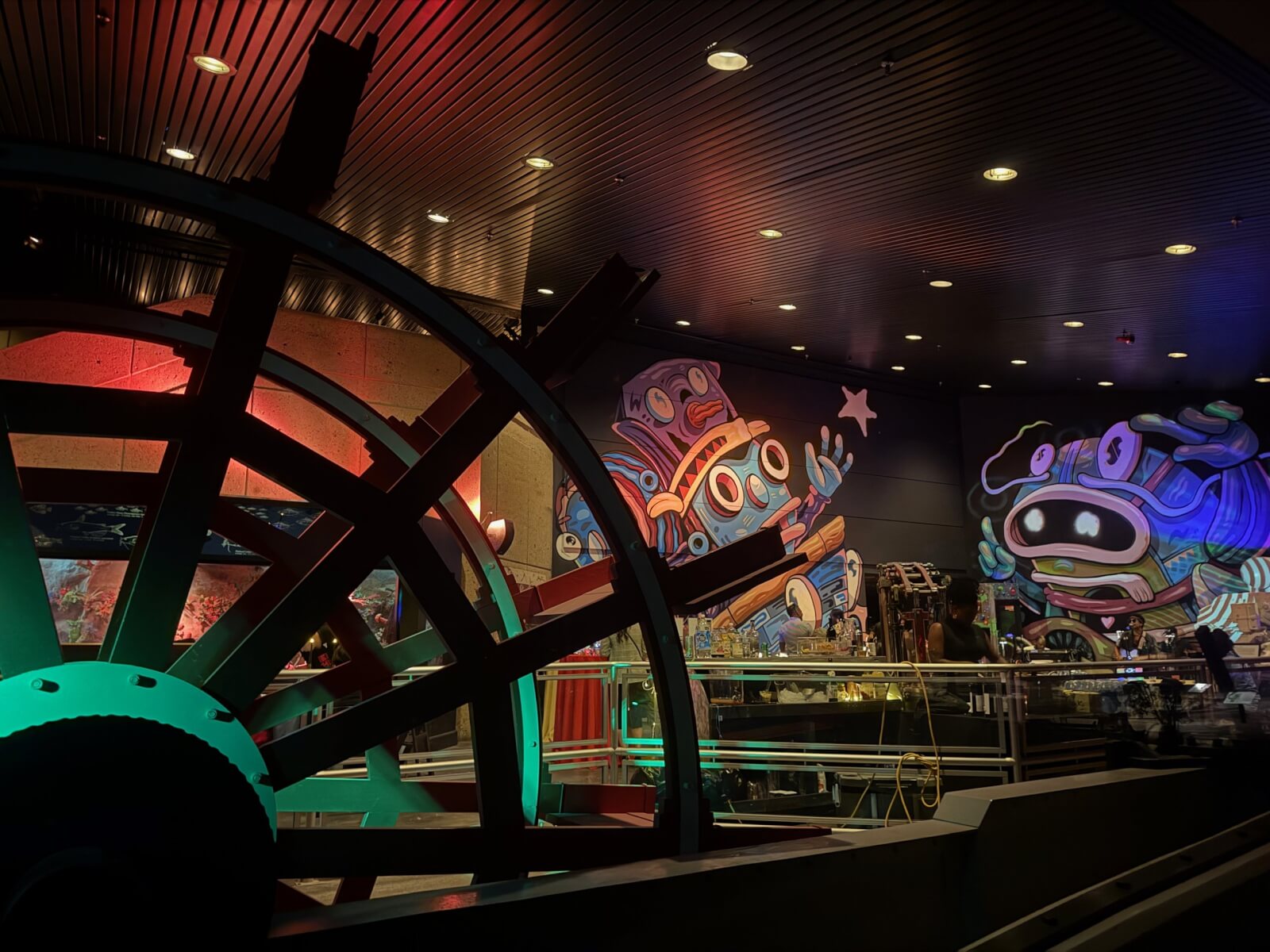 A Sneak Peek at Memphis's Most Buzzed-About New Experience - 7 Large water wheel in foreground with vibrant, cartoonish murals of robots on the back wall, setting the stage for an indoor adventure that captivates all ages under bright artificial lighting.