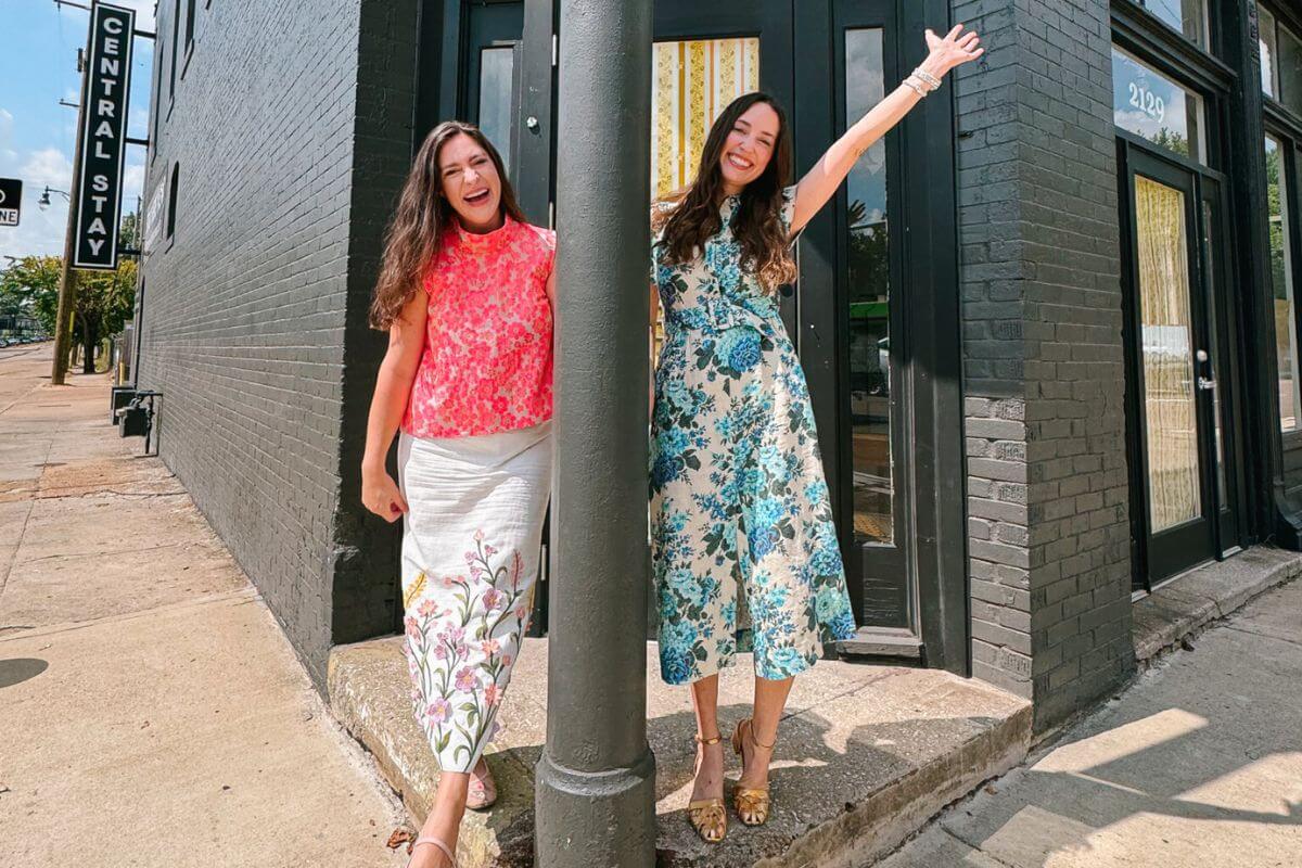 Dorothy Collier and Alice Kerley, both in floral dresses, stand on a city sidewalk by a black lamppost, smiling and posing in front of a dark brick building.