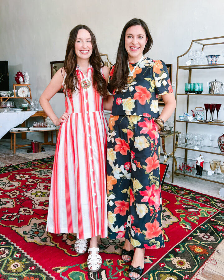 Meet the Owners of Memphis's New Elevated Vintage Shop: Heirloom House - 2 Two women stand on a colorful rug; one wears a red and white striped dress, the other wears a navy floral jumpsuit. Shelves with glassware and decor are in the background.