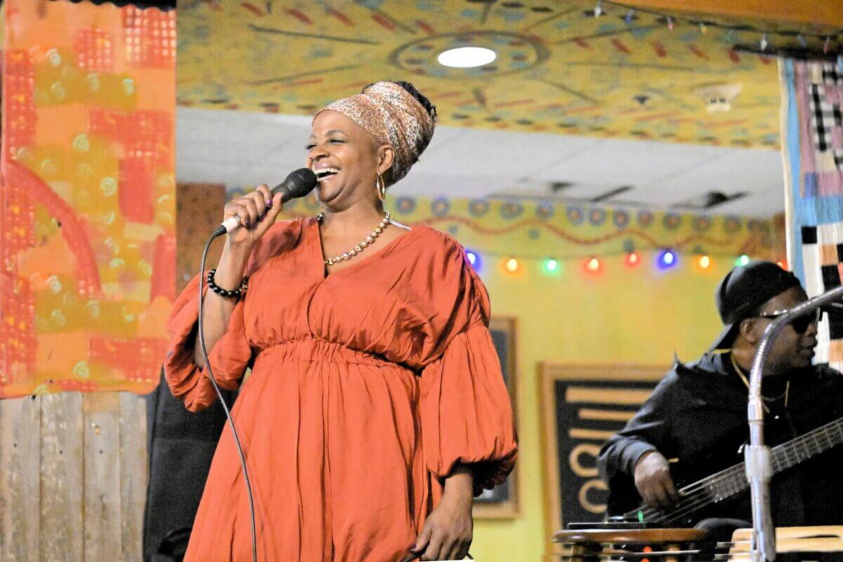 Cequita Monique, in an orange dress, sings into a microphone onstage, smiling as the bassist plays in the background amidst colorful FACES decorations.