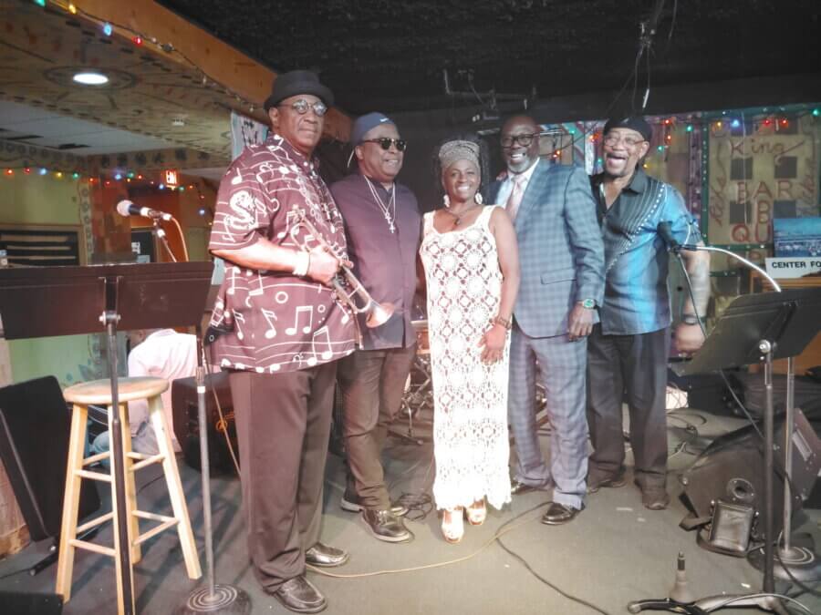 Meet Memphis Jazz Musician Cequita Monique - 6 Five musicians, including Cequita Monique, stand together on stage as FACES, smiling at the camera. Musical instruments and microphones surround them, with colorful lights glowing in the background.