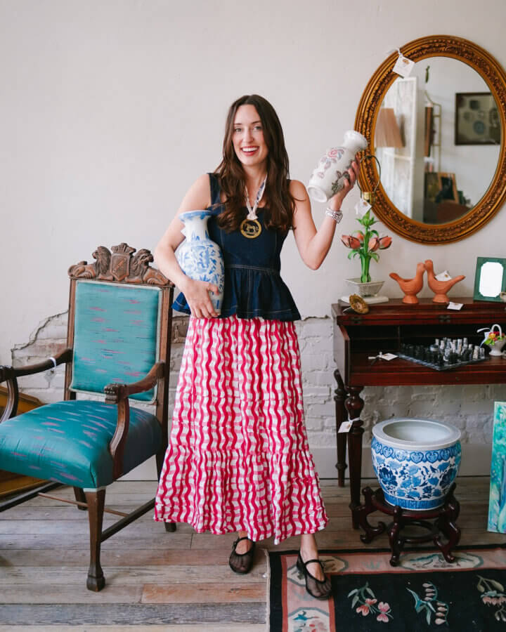 Meet the Owners of Memphis's New Elevated Vintage Shop: Heirloom House - 4 Woman standing indoors holding two vases, wearing a sleeveless navy top and a long red patterned skirt, surrounded by furniture and decorative items from Dorothy Collierβs and Alice Kerleyβs collections.