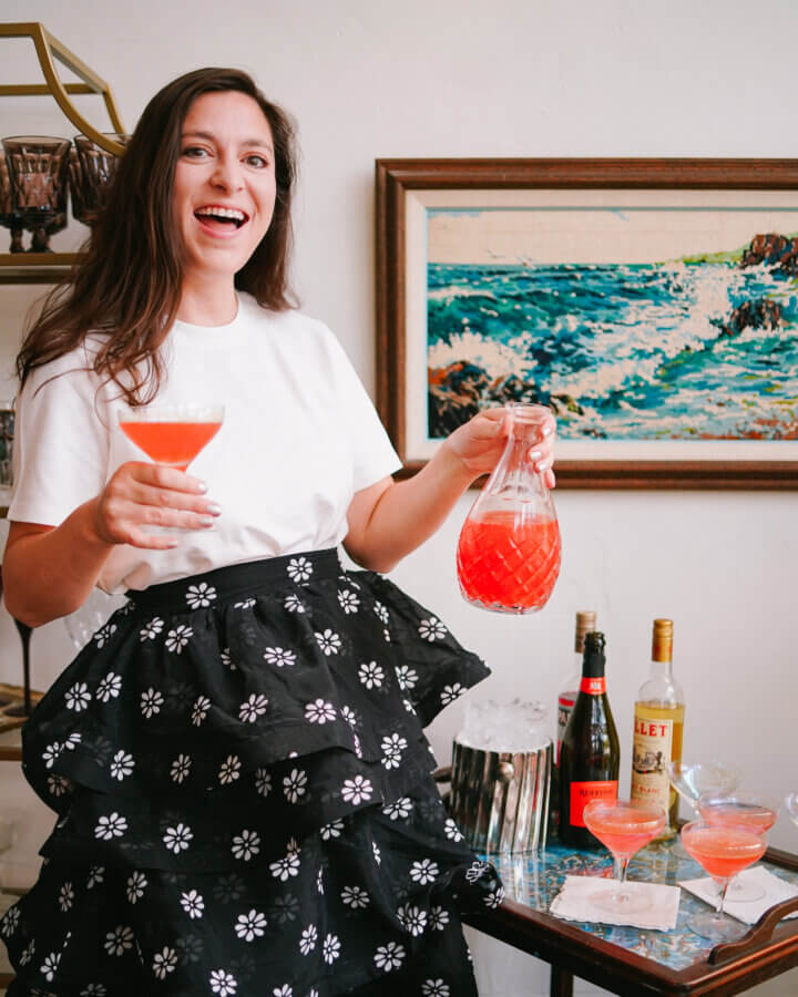 Meet the Owners of Memphis's New Elevated Vintage Shop: Heirloom House - 5 A woman in a white shirt and black floral skirt holds a carafe and glass of pink drink, standing by a table with bottles and cocktails in front of a Dorothy Collier landscape painting.
