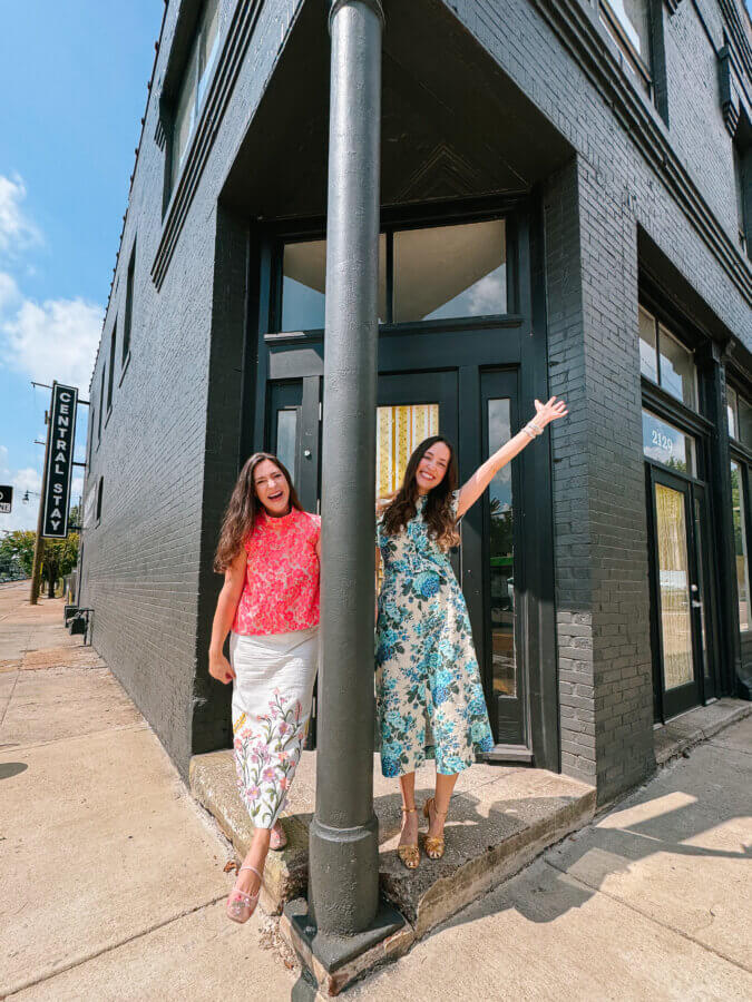 Meet the Owners of Memphis's New Elevated Vintage Shop: Heirloom House - 3 Alice Kerley and Dorothy Collier stand on a small corner step outside a black brick building, smiling for FACES; one holds onto a pole while the other raises an arm, both dressed in colorful summer outfits.