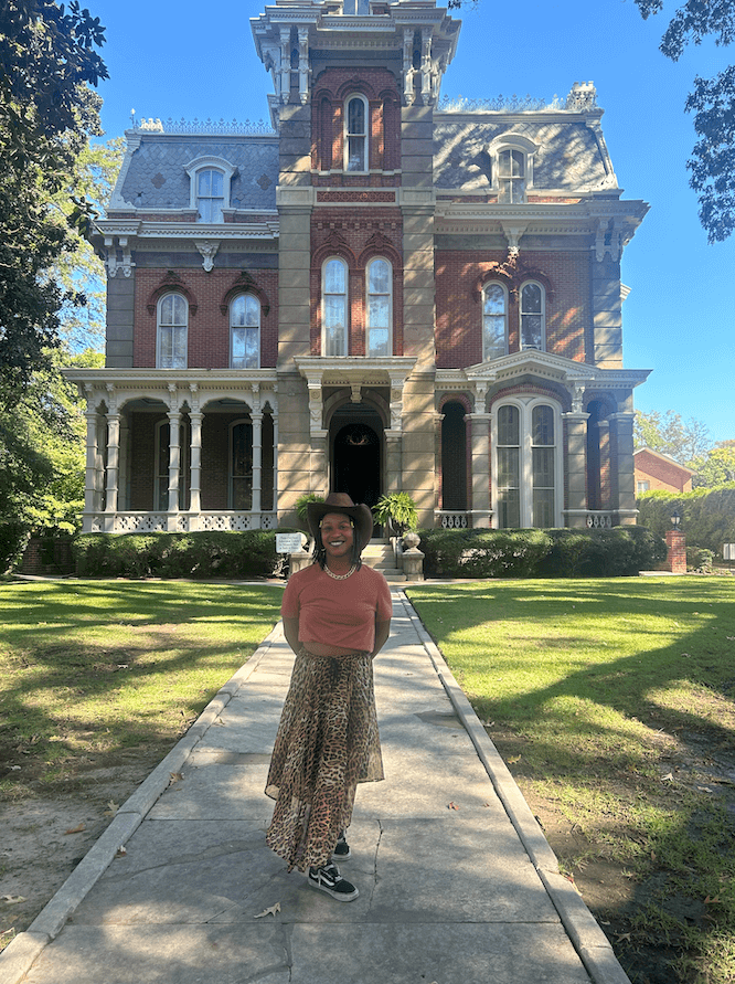 Meet the Woman Behind the New Memphis Rap Tour - 6 A woman, Taylor Nicole Barksdale, stands smiling on a walkway in front of a large, ornate Victorian-style brick house on a sunny day.