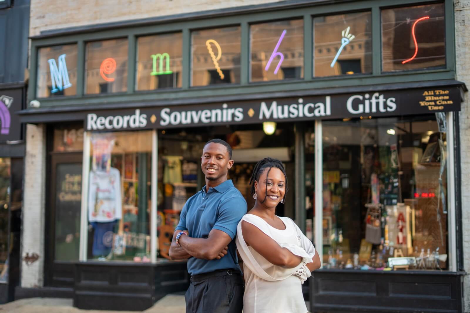 Meet the Woman Behind the New Memphis Rap Tour - 4 Two people stand back-to-back, smiling with arms crossed, in front of a storefront displaying “Memphis” and signs for records, souvenirs, and musical gifts.
